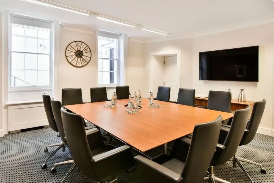 A meeting room with a wooden table and black chairs in front of a window and a clock.