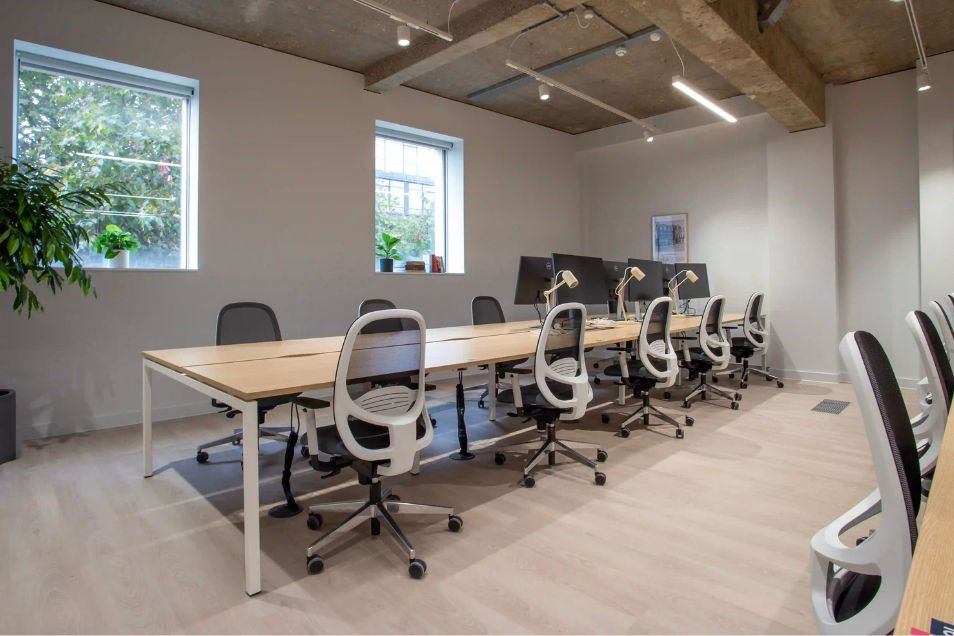 Rows of wooden desks and black chairs in an office with windows and exposed ceiling beams.