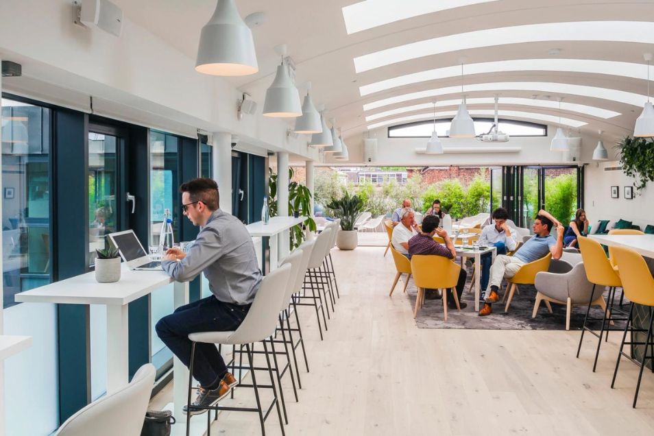 People at tables and a counter working on laptops in a communal area with plants and arched windows.