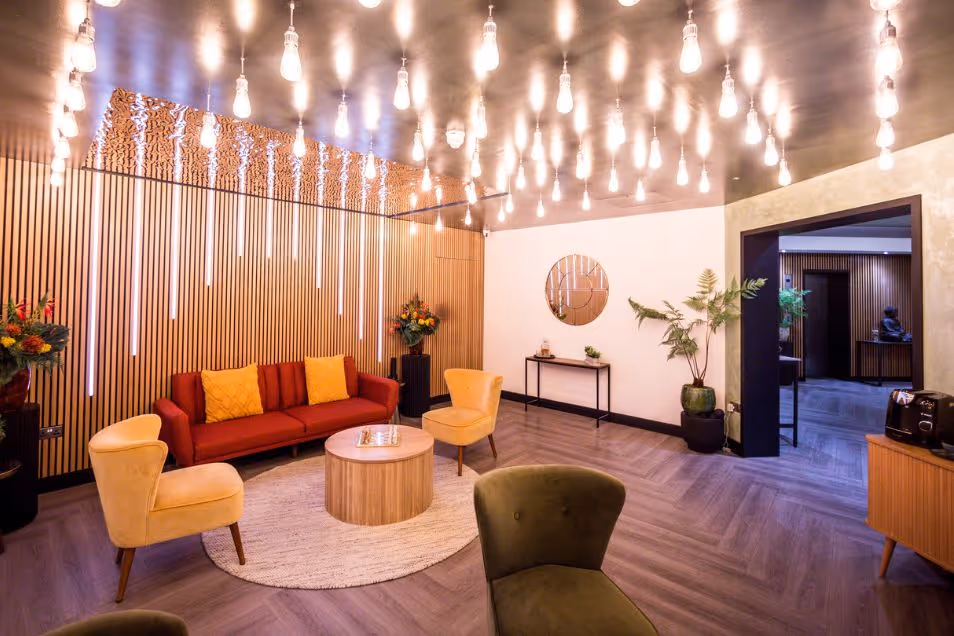 Armchairs and a sofa surround a circular table in a lobby with wood floors and lights hanging from the ceiling.