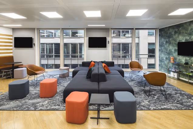 A lounge features sofas and stools on a rug in an office with windows and television screens.