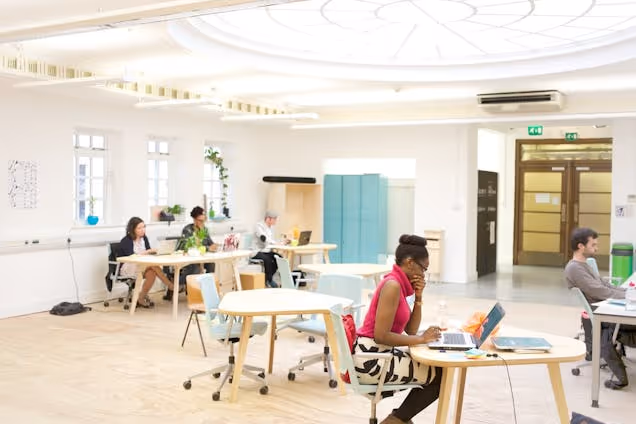 People sit at wooden desks, working on computers in a white office with a circular roof lantern and blue cabinets.