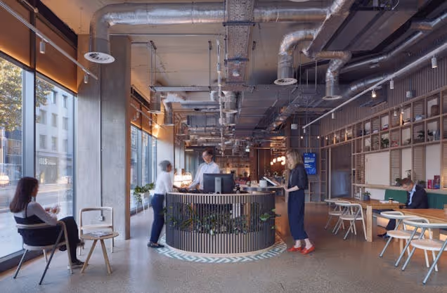 People talk at a circular reception desk in an industrial office space with exposed ceiling pipes.