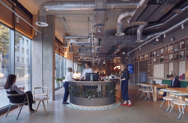 People talk at a circular reception desk in an industrial office space with exposed ceiling pipes.