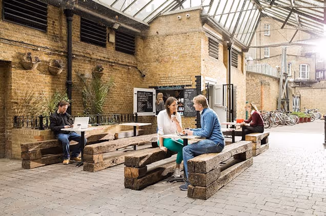 People sat at wooden tables talking or working on laptops in an outdoor courtyard with brick walls.