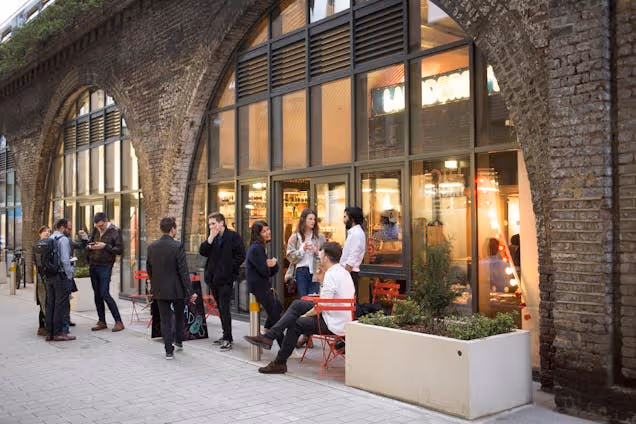 People stand and talk outside a building with glass doors and a white planter under a brick archway.