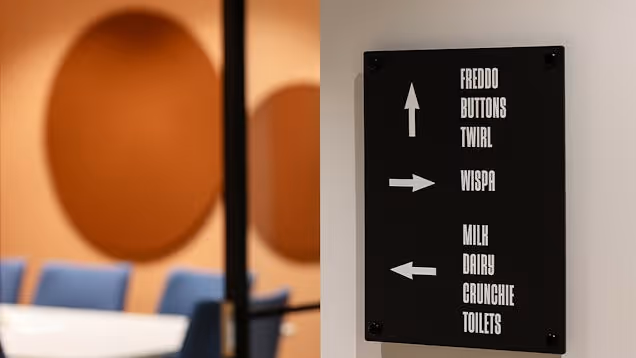 A black sign with white text and arrows points toward meeting rooms and toilets next to a glass wall that looks into a meeting room that has a white table, blue chairs and an orange wall.