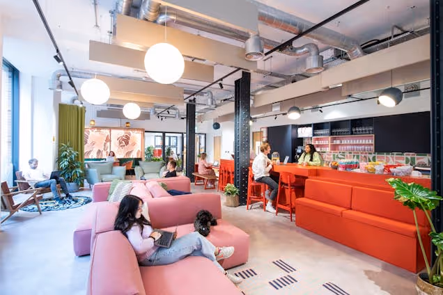 People on pink sofas and at an orange kitchen island in an office lounge with globe lights and ceiling pipes.