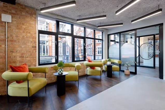Green armchairs and small black tables sit in an office breakout area with brick walls and large windows.