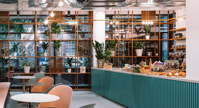 Chairs and circular tables sit in front of an office cafe counter with shelves holding plants and food items.