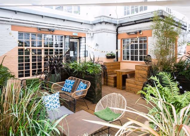 Chairs and wooden tables sit on a roof terrace surrounded by lots of plants and sunlight.
