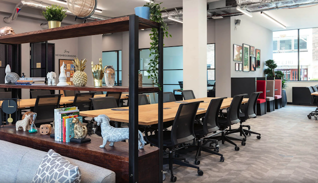 Black chairs at a wooden table in an office with shelving, coworking booths, and plants.