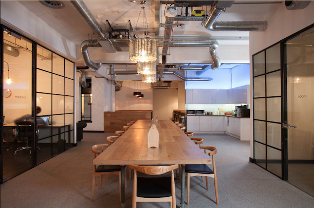 A wooden table with chairs in a communal space with a kitchen and glass partitions.