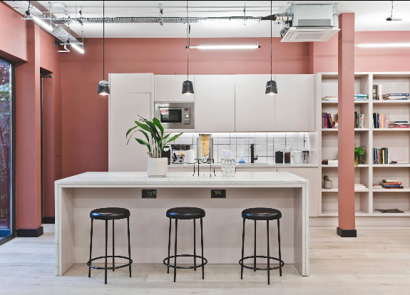 Three black stools sit at a white kitchen island in an office with pink walls and shelves.