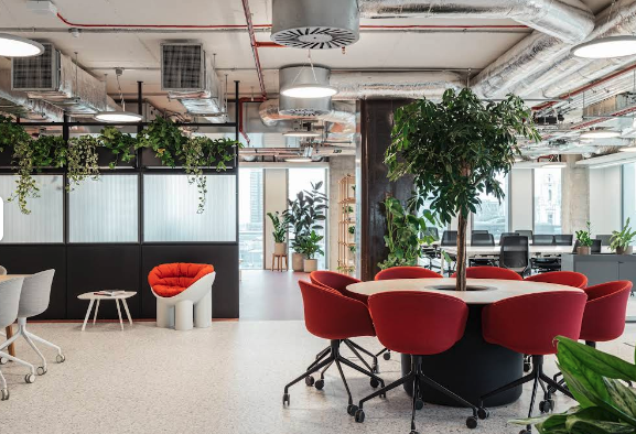 Red chairs surround a wooden table with a tree in the centre inside a coworking office.