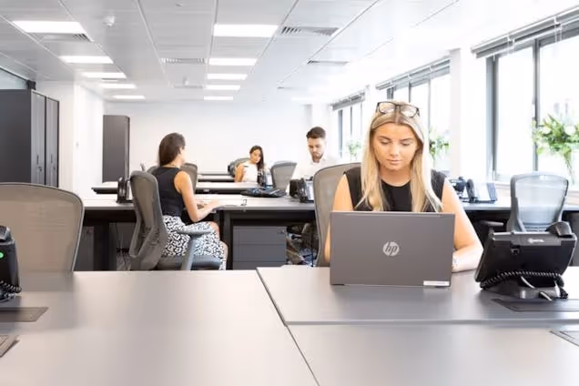 People sitting at desks working on laptops in an office with windows and grey dividers.