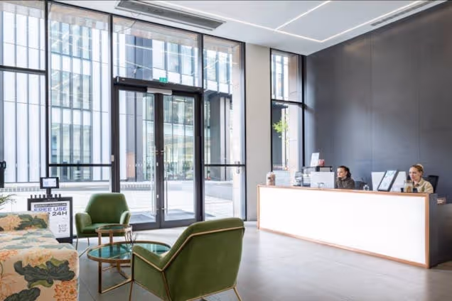 A reception desk with workers behind it in an office lobby with green chairs and windows.