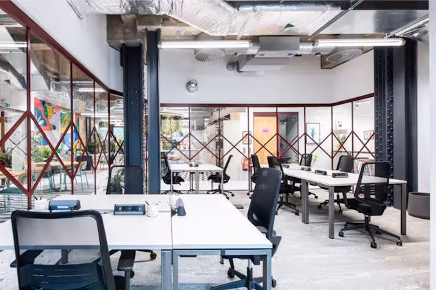 An office space with white desks and black chairs arranged in rows under exposed ceiling pipes.