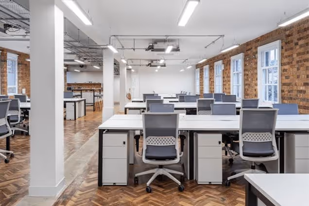 Rows of white desks and black mesh chairs sit on a parquet floor in an office with brick walls.