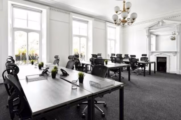 Rows of desks with office chairs and monitors sit in a room with a chandelier, fireplace, and large windows.