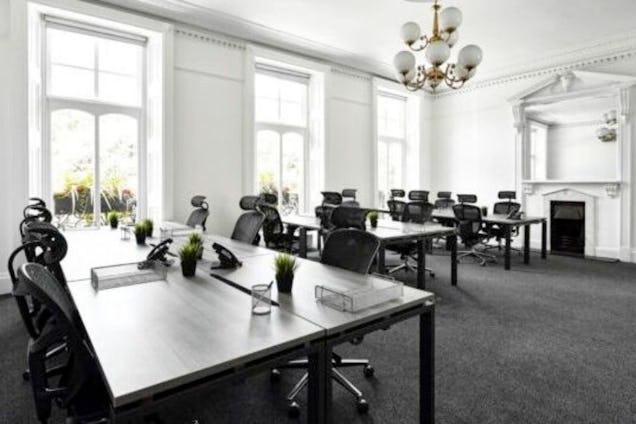 Rows of desks with office chairs and monitors sit in a room with a chandelier, fireplace, and large windows.