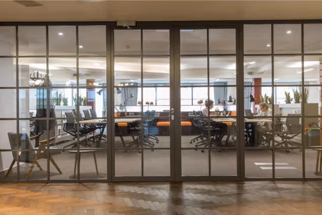 An office with desks and chairs is visible through glass partition doors on a parquet floor while a person sits at a desk.