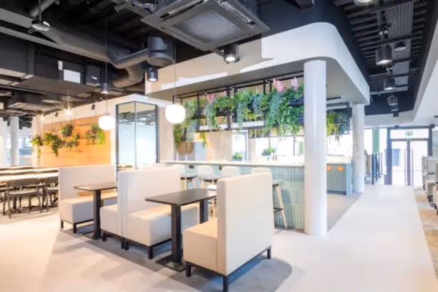 Rows of booths and tables sit in a coworking area with hanging plants and exposed ceiling pipes.