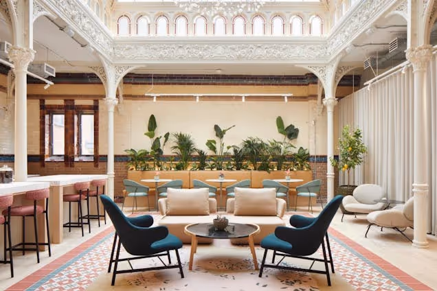 Teal and cream chairs and a coffee table sit in a lounge with plants and pillars under a skylight.