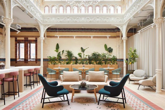 Teal and cream chairs and a coffee table sit in a lounge with plants and pillars under a skylight.