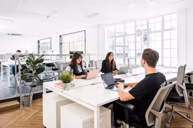 People at white desks using laptops in a bright office with large windows and plants.