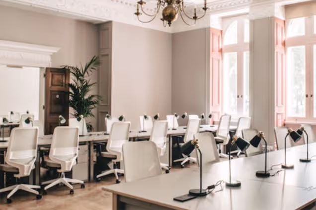 White long chairs at light-coloured desks in an office with large windows, lamps at each desk, a fireplace, and a chandelier.