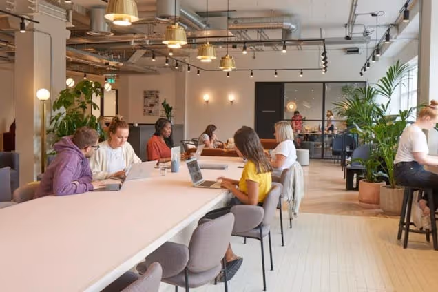 People sat, working at a long pink table in a coworking space with scattered plants and sphere lights.