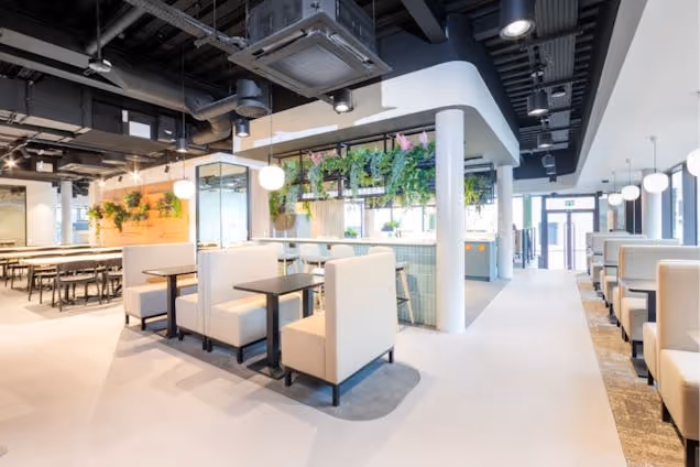Rows of booths with tables sit in an office coworking area with hanging plants and exposed ceiling pipes.