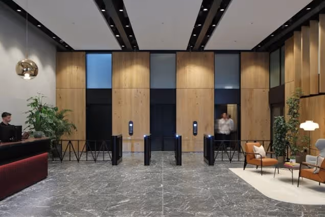 Security turnstiles stand in a building reception area with marble floors, wood wall panels, and a person standing in the background.