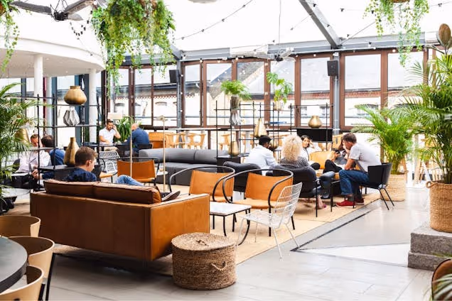 People sat on tan sofas and black chairs in a communal area with plants under a glass roof and windows.
