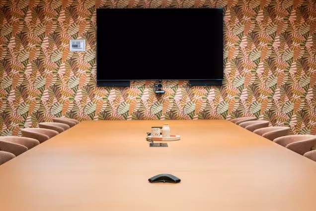A tan wooden table and pink chairs in a boardroom with a black television on a green and pink patterned wall.