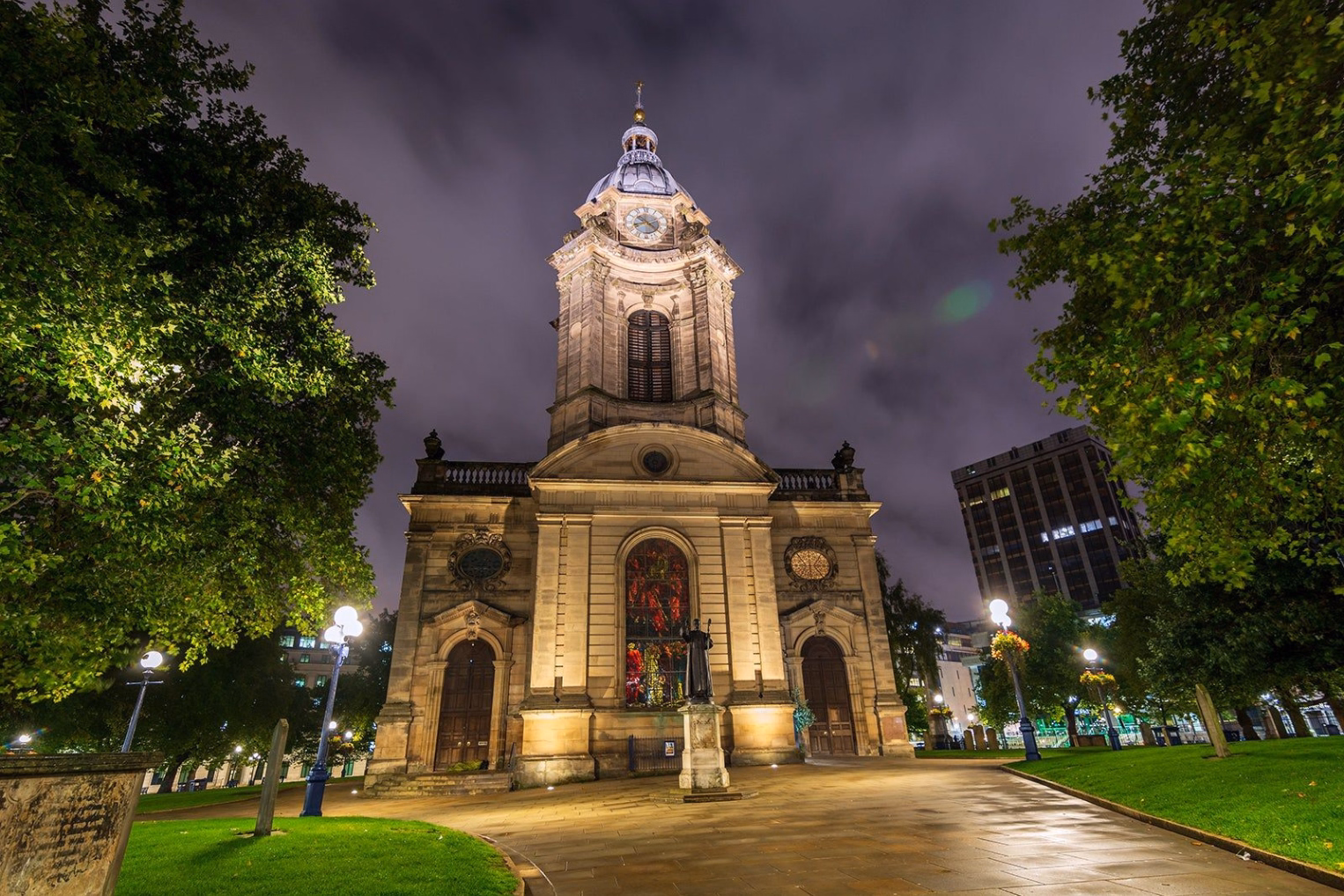 St. Philip's Cathedral in Birmingham illuminated at night, featuring a stone tower and clock face surrounded by green lawns and trees.