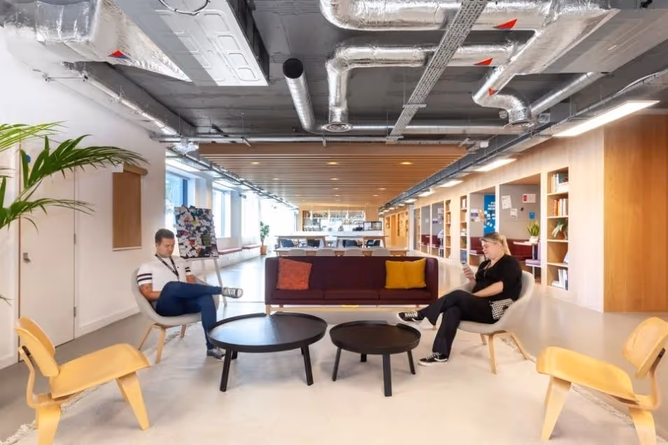 Open-plan office lounge with exposed industrial piping, featuring two people sitting in grey armchairs near a burgundy sofa and round black coffee tables.