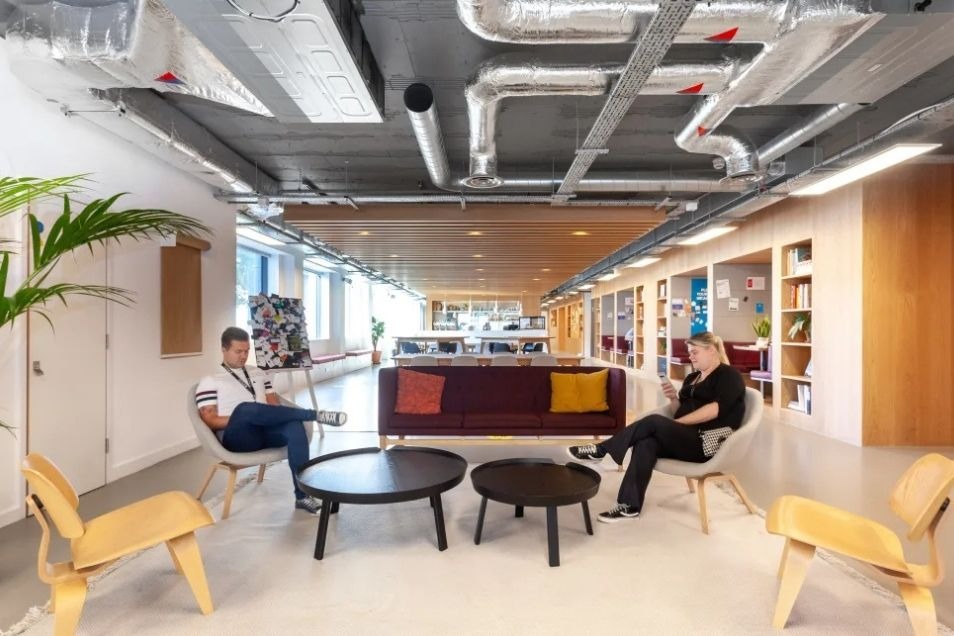 Open-plan office lounge with exposed industrial piping, featuring two people sitting in grey armchairs near a burgundy sofa and round black coffee tables.
