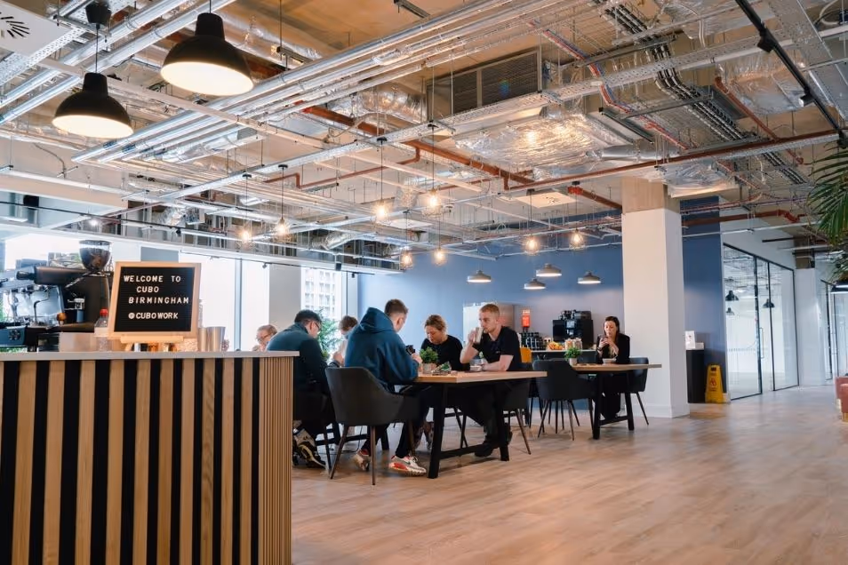 Spacious office cafe with exposed industrial ceiling pipes and pendant lights, featuring people seated at a long wooden table. 