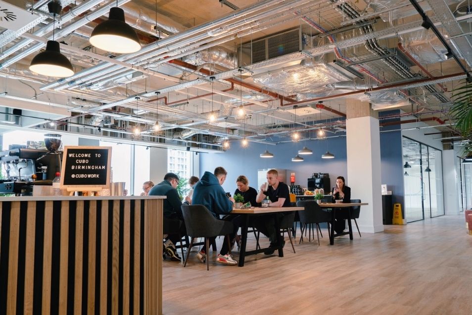 Spacious office cafe with exposed industrial ceiling pipes and pendant lights, featuring people seated at a long wooden table. 