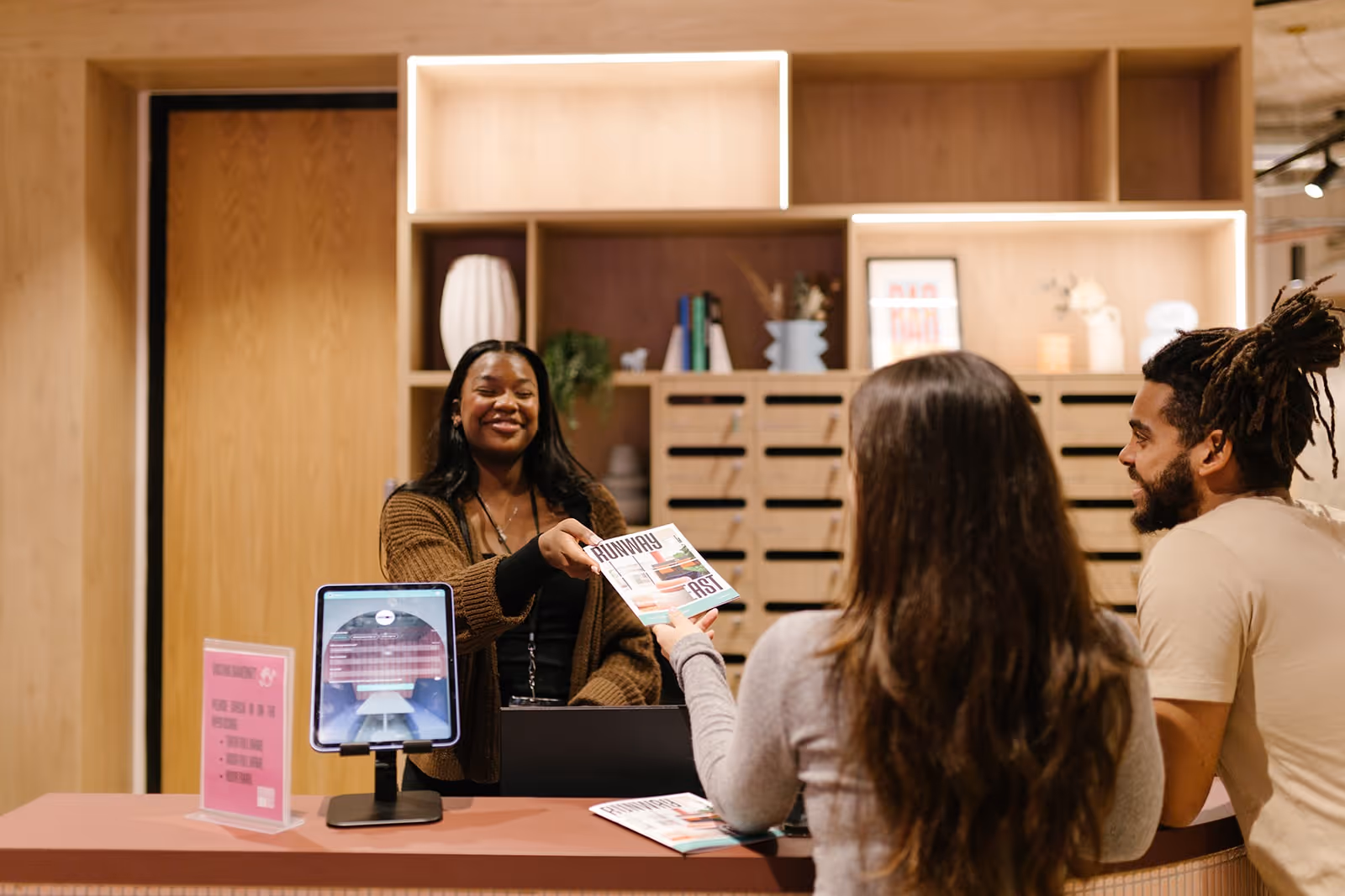 A receptionist smiles while handing a pamphlet to two people at a wooden front desk in a well-lit office reception area.