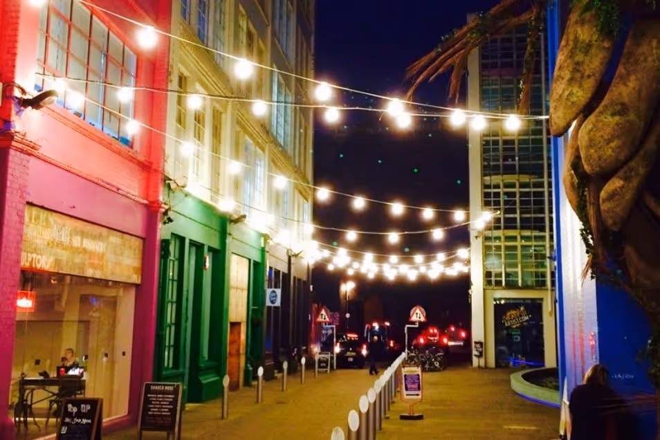 A vibrant street at night, lined with colourful buildings in pink, green, and blue, illuminated by strings of overhead globe lights.