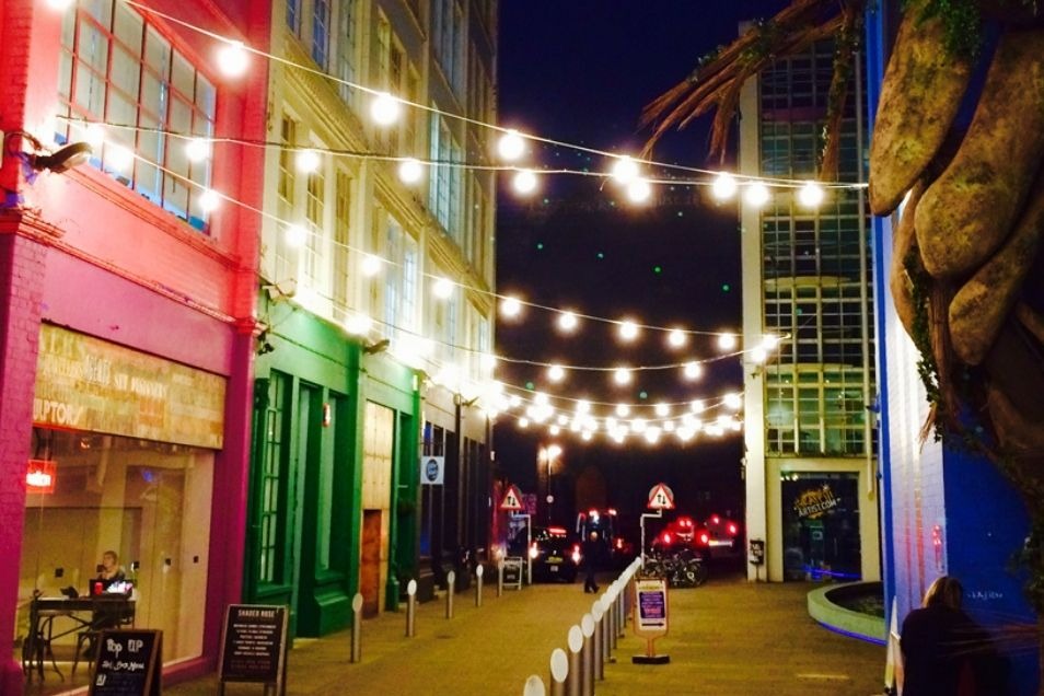 A vibrant street at night, lined with colourful buildings in pink, green, and blue, illuminated by strings of overhead globe lights.