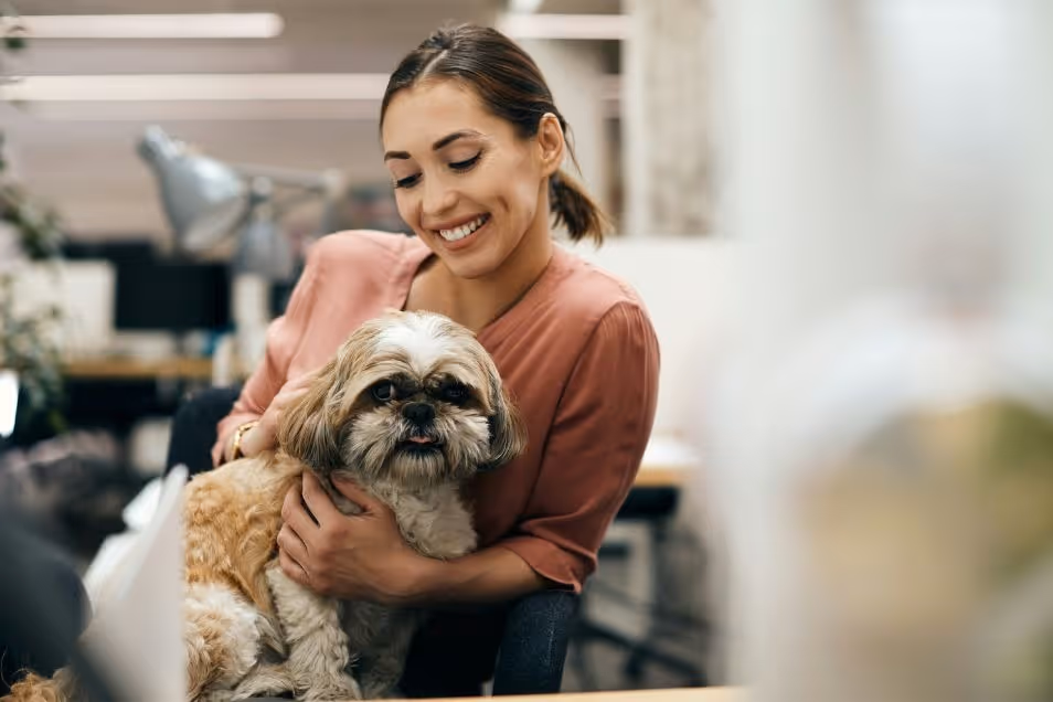 A smiling woman in a pink top sits in a bright office environment while hugging a fluffy dog.