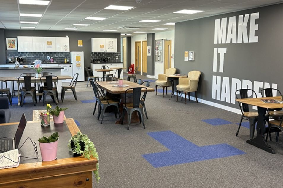 An open-plan office breakroom with long communal tables, a coffee station, and a "MAKE IT HAPPEN" wall graphic.