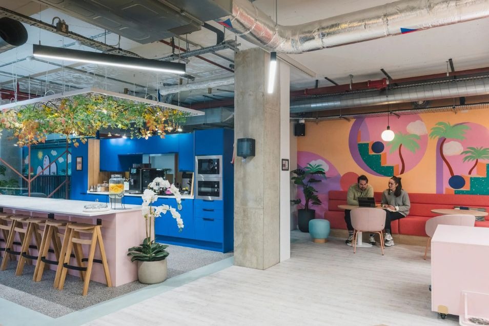 Office kitchen and cafe area where two people sit and talk against a colourful mural wall.
