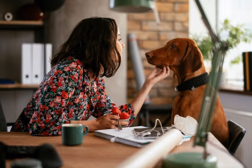 A woman in a floral shirt sits at a desk and affectionately pets a brown dog in a home office setting.