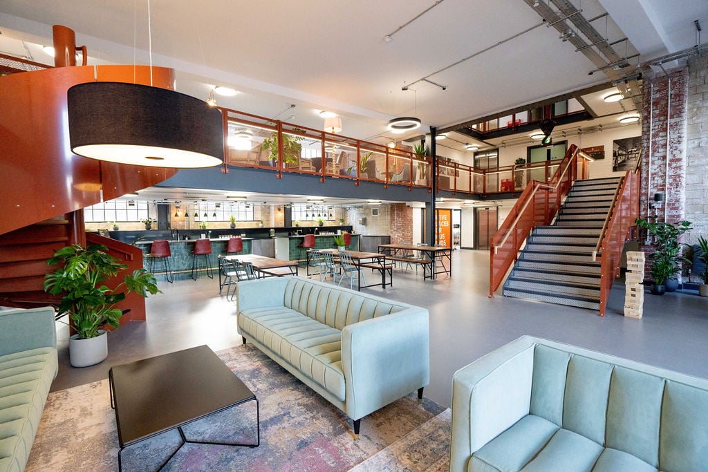 A spacious multi-level office lounge featuring a spiral staircase, light blue tufted sofas, and a wooden mezzanine.
