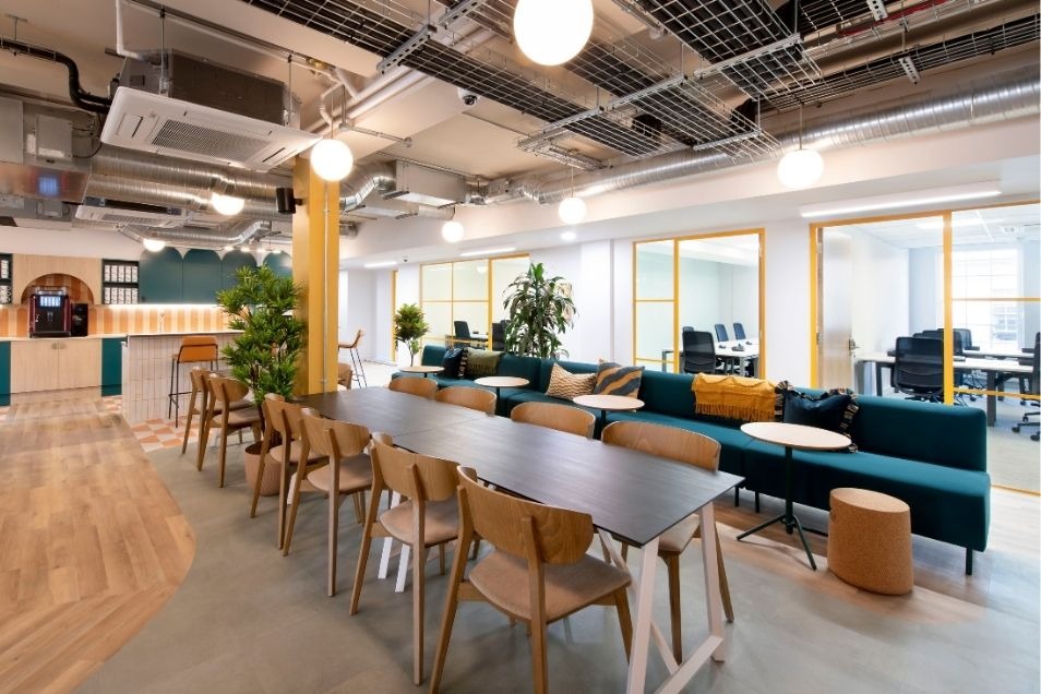 Office break room with a long communal table, wooden chairs, and a deep teal sofa under industrial lighting.